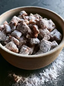 Close-up high-angle shot of puppy chow recipe in a ceramic bowl, featuring velvety powdered sugar and gooey chocolate underneath.