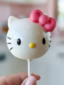 A handheld smartphone photo showing a flawlessly smooth white chocolate hello kitty cake pop against a pastel kitchen background. (hello kitty cake pops)