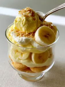 A handheld smartphone photo showing a silver spoon scooping into a cup of banana pudding with distinct layers of wafers and fruit. (banana pudding)