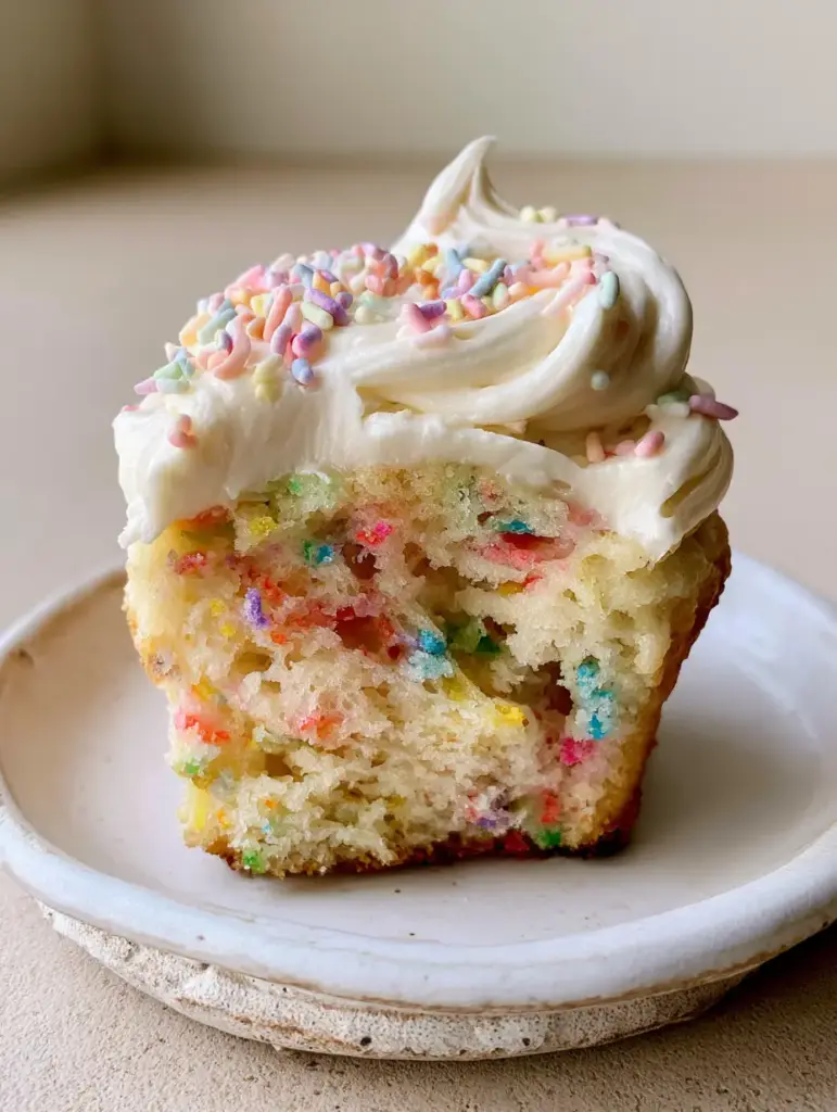 Eye-level shot of a single funfetti cupcake with a silky vanilla buttercream swirl and pastel rainbow jimmies on a rustic plate.