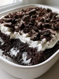 A high-angle smartphone photo of an Oreo Poke Cake in a matte white ceramic dish, covered in whipped cream and jagged cookie pieces under natural morning light.